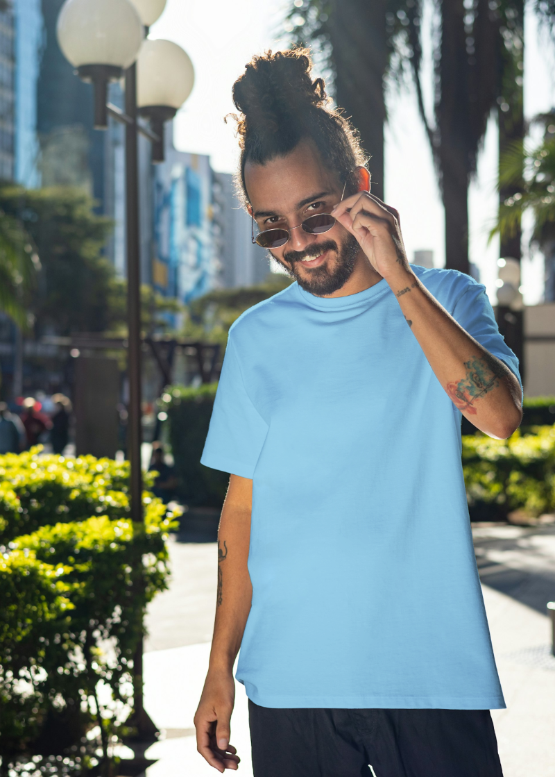 Man in a light blue t-shirt talking on a phone outdoors with greenery and buildings in the background