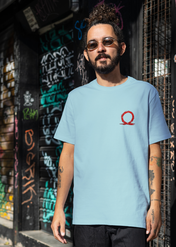 Man wearing a light blue t-shirt with a red logo in front of a graffiti-covered wall.