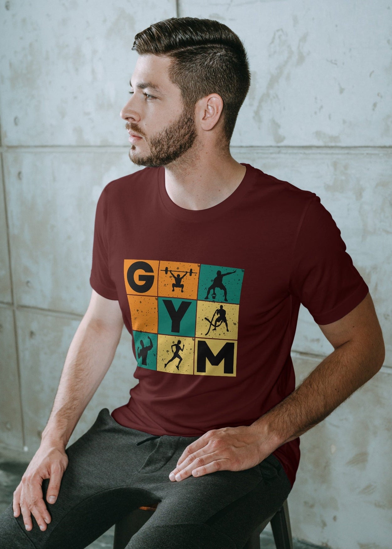 Man wearing a maroon t-shirt with a colorful 'GYM' graphic design sitting against a textured wall.