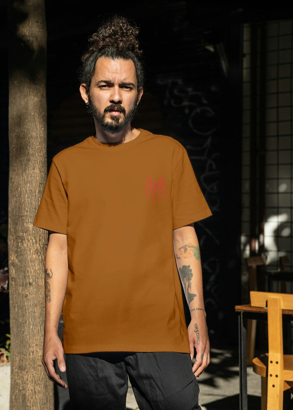 Man wearing a brown t-shirt standing outdoors with plants and tables in the background